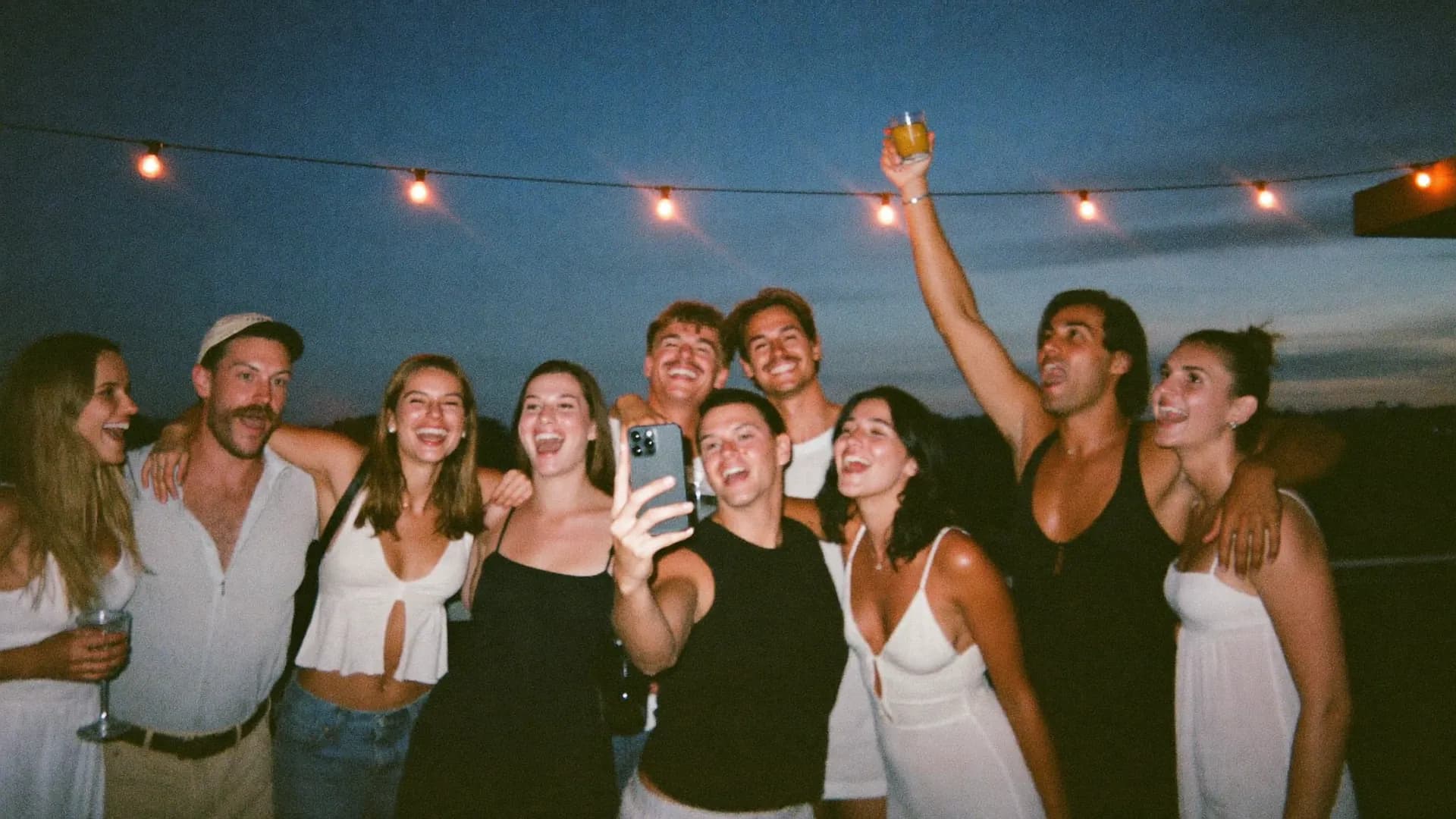 Neun Freundinnen und Freunde stehen abends unter Lichterketten zusammen und machen lachend ein Gruppenfoto