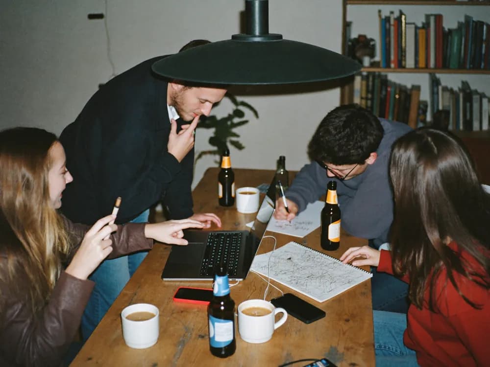 Vier Freunde sitzen abends mit Laptop, Handys und Notizblock um einen Küchentisch und planen
